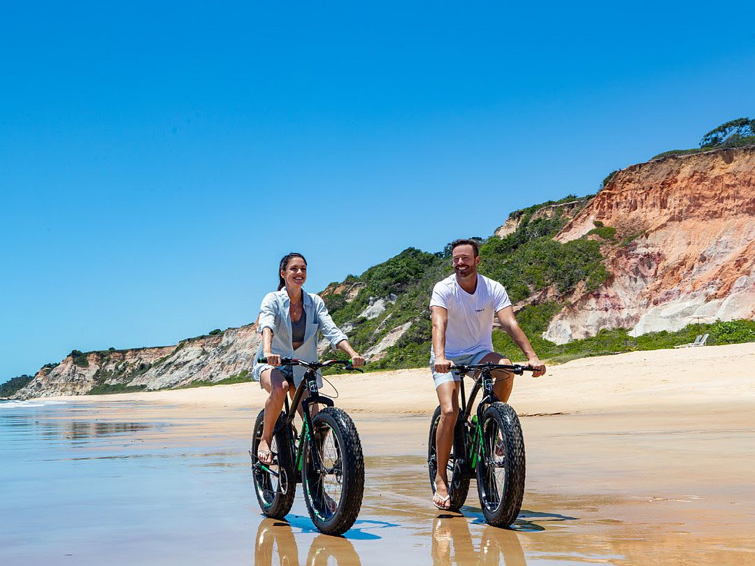 Casal em fat bikes na praia, com o mar e as falésias ao fundo